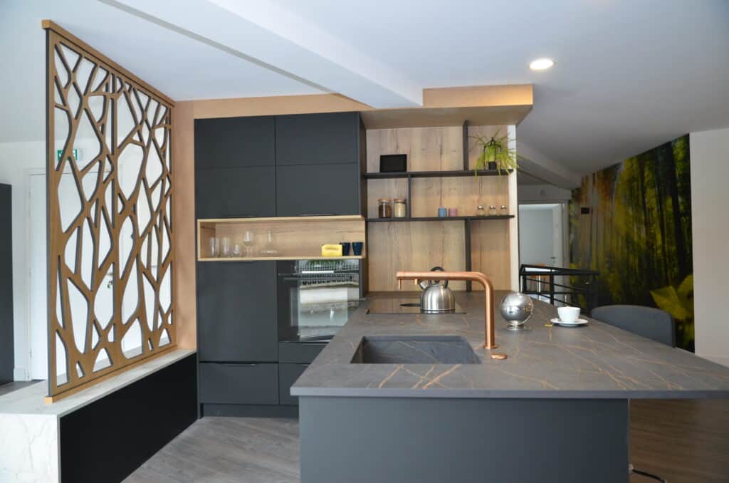Cuisine design avec un îlot central en pierre noire veinée, une robinetterie en cuivre, des façades noires mates, des étagères en bois clair, et une cloison décorative ajourée en bois ajoutant une touche moderne et élégante