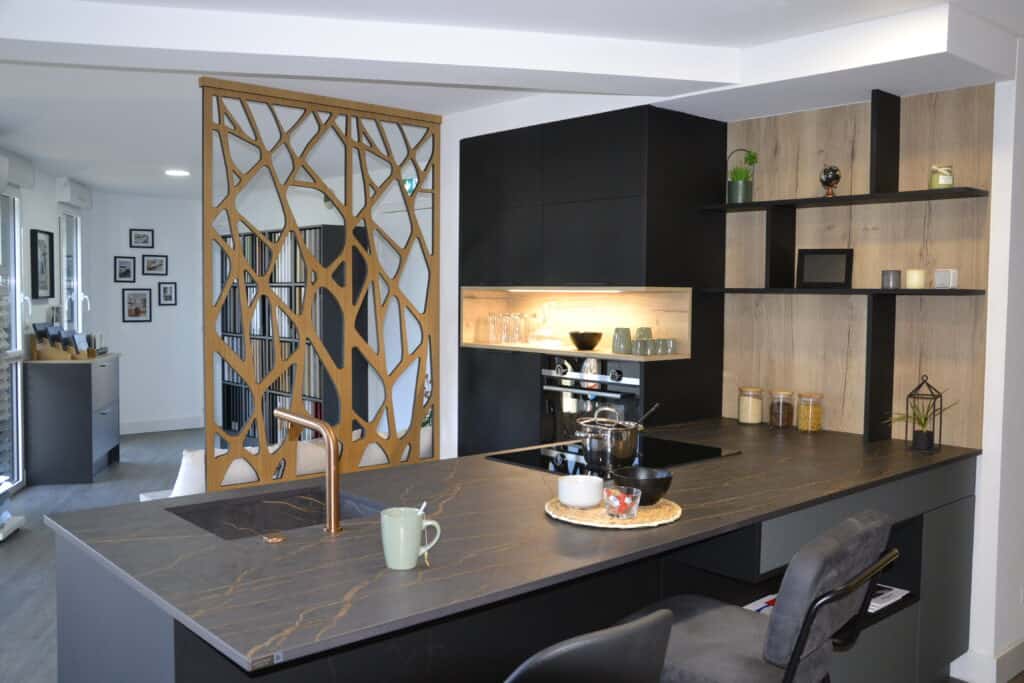 Cuisine moderne et élégante avec un îlot central en pierre noire veinée, une robinetterie en cuivre, des rangements en bois et noir mat, ainsi qu’une cloison décorative ajourée en bois, créant un espace à la fois fonctionnel et esthétique.