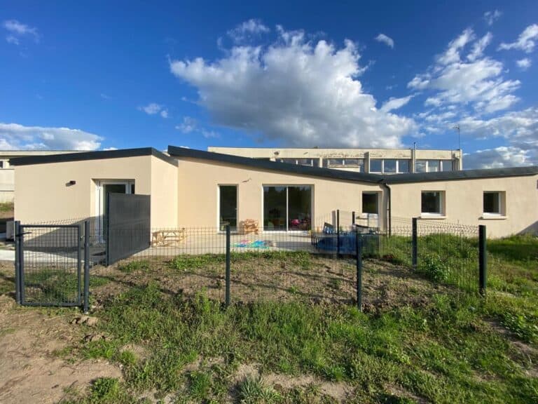 Photo de la façade d’un bâtiment récemment rénové, avec un crépi beige uniforme et une toiture en pente moderne. Un jardin clôturé entoure l’espace extérieur, et de grandes baies vitrées permettent une luminosité optimale. Le ciel bleu et dégagé met en valeur les finitions impeccables de la structure