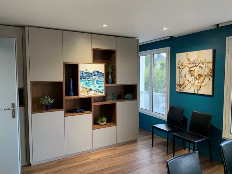 Un espace élégant et organisé, mettant en valeur un meuble sur mesure combinant rangement fermé et étagères ouvertes en bois. La profondeur des niches met en avant des objets décoratifs et des œuvres d’art, comme le tableau central aux teintes bleutées. Le mur bleu profond apporte une ambiance chaleureuse et moderne, accentuée par des touches de lumière naturelle provenant des grandes fenêtres. Le sol en bois ajoute une note de confort et de sophistication à l’ensemble.